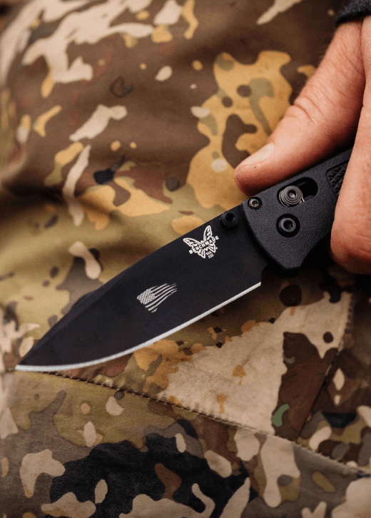 Custom black Benchmade Bugout folding knife with a company logo laser-marked on the blade, shown against camouflage workwear.